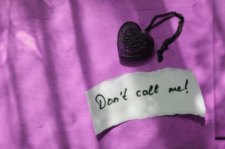 Handwritten note Don't Call Me in a white piece of paper and a wooden amulet in the shape of a heart. Lilac background, shadows. Love, Relationship Concept. Top viewの写真素材