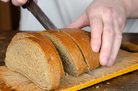 Hands slice bread on a wooden cutting board. A grown man slices a loaf of rye bread with a large...