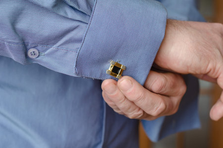 Close-up of hands buttoning a cufflink on the sleeve of a blue shirt. A middle-aged adult man with a...