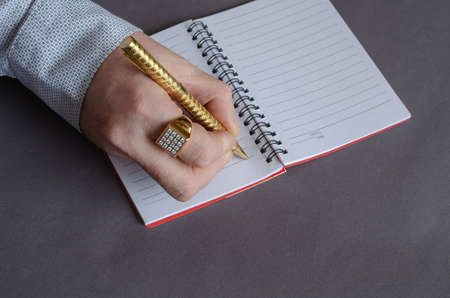 A hand writing with a gold pen in an open notebook on a spring. A grown man with a seal on his index finger. Business concept. Gray background. View from above at an angle.の写真素材