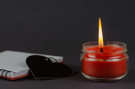 Red candle, notepad and black heart symbol on gray background. Burning wax candle in a glass jar. Open, blank, spring-loaded notebook. Side view at an angle. Selective focus.の写真素材