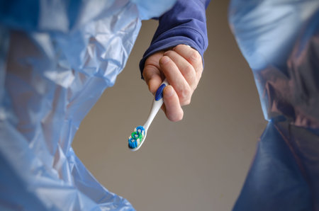 Hand tosses an old plastic toothbrush into the garbage can. An adult male holds a toothbrushing tool...