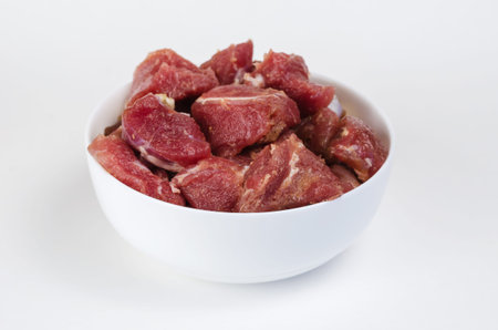 Ceramic bowl with raw marinated meat on a white background. Pieces of pork with salt, pepper and...