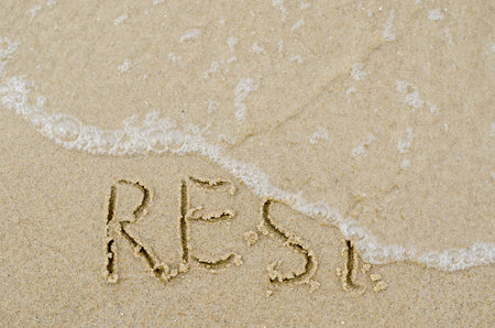 The wave washes away the word REST in the yellow sand. Handwritten word on wet sandy beach. Capital...