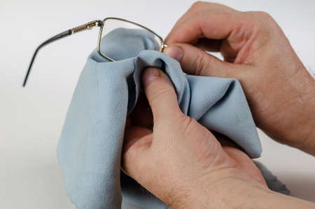 Hands holding reading glasses and wipe on white background. A man is cleaning his metal-rimmed glasses with a special blue cleaning cloth. Close-up. Selective focus.の写真素材