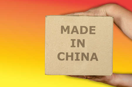 Hands holding a cardboard box on a colored background. A man with a square package of recycled material. Inscription made in China. Global trade concept. Side view.の写真素材
