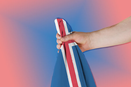 National flag Kingdom of Iceland in hand on red and blue background. Man holding flag of country located on an island in northern Europe. Side view.の写真素材
