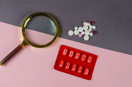 Magnifying glass and medical pills on a gray-pink background. A group of random pills and pills. Searching for the right medicine concept. Top view. Copy space.の写真素材