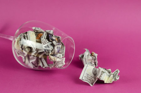 Transparent wine glass with money lying on pink background. Glass is full Crumpled hundred dollar bills.の写真素材