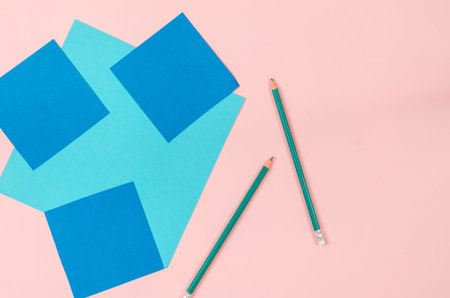 Sheets of colored paper and green pencils on a pink background. Square blue and light blue sheets....