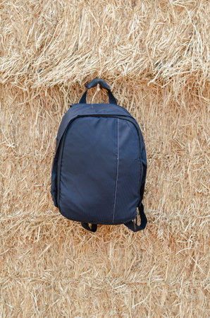 The backpack hangs in front of the hay depot. A black casual satchel. A wall of yellow dried grass....