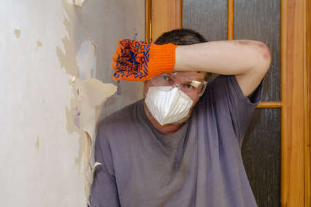 An adult male is wiping sweat from his forehead leaning against the wall. Tired male in respirator, goggles, orange gloves. Repair, Removal of paper wallpaper. Inside the room. Series part.の写真素材