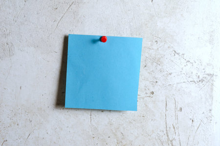 Blue square piece of paper. The note paper is attached with a stationery button. A gray cement wall with scratches. Vertical.の写真素材