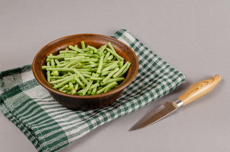 Brown clay bowl with raw green beans. Kitchen knife with wooden handle. White and green checkered cloth. Healthy food. Close-up. Selective focus.の写真素材
