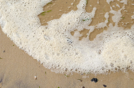 Wave with sea foam on the sandy shore. Sea water with green algae. Yellow sandy beach. View from above at an angle. Selective focus, blurred motion, noise, grain effect.の写真素材