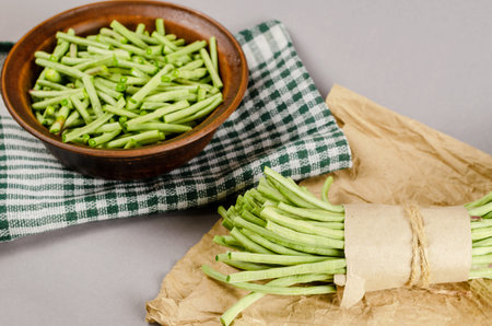 A bundle of raw green beans. Brown bowl of sliced bean asparagus. A flocked cloth. Gray background....