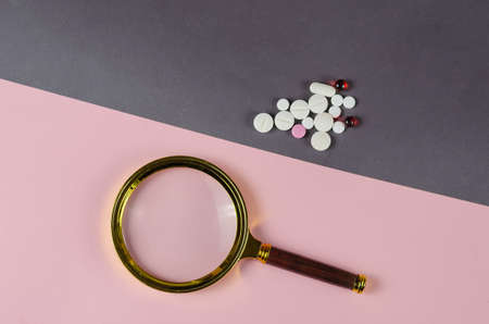 Magnifying glass and medical pills on a gray-pink background. A group of random pills and tablets with no packaging. Searching for the right medicine concept. Top view. Copy space.の写真素材