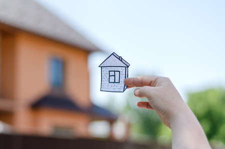 Woman holding a drawing of a house in front of a blurred cottage. Woman's hand showing a paper figure of a house. Defocused red cottage or townhouse building. Real estate, rentals, sales.の写真素材