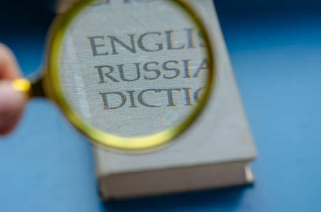 Education, foreign language learning, search for knowledge concept. Magnifying glass and hardcover book on blue background. English-Russian dictionary enlarged in the loupe.の写真素材