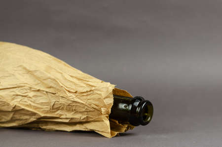 Drunken Bottle in paper bag lies on gray background. Dark empty bottle of alcohol in crumpled brown bag. Social problem of alcoholism concept. Close-up. Selective focus.の写真素材