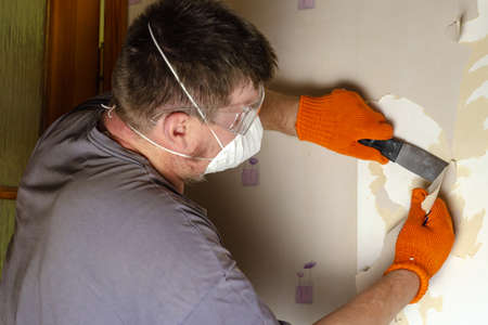 An adult man removes old wallpaper from the wall. The male is wearing a respirator, safety eyeglasses, and orange gloves. Dismantling paper wallpaper. Inside the room. Series part. Selective focus.の写真素材