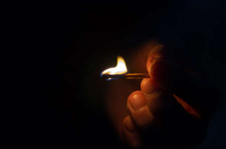A mans hand with a burning match against a black background. Close-up of a mans hand with a match lit in the night. Defocus, noise, blurred, grain effect.の写真素材