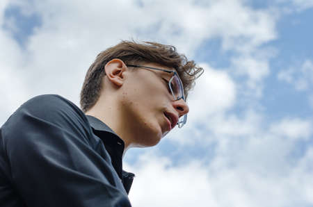 A portrait of a young adult male against a blue sky. A man wearing a black shirt and optical glasses. View from bottom to top. Selective focus.の写真素材