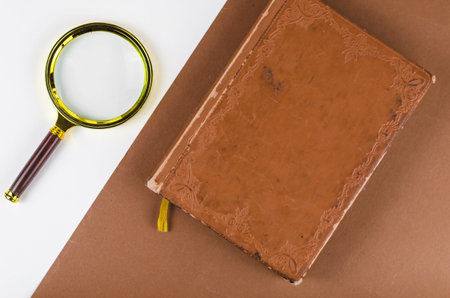 Magnifying glass and an old notebook on a two-color background. A diary or planner with notes in brown leather binding. Minimalist composition on white and brown. Top view. Copy spaceの写真素材