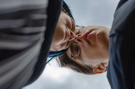 A heterosexual couple in love kissing against the sky. A young man wearing optical glasses. Young woman with braided thin pigtails. View from bottom to top. Selective focus.の写真素材
