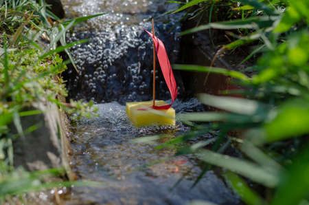 A yellow childrens boat with a red sail into a narrow channel or creek. A small sailboat floats down a narrow stream among green grasses. Blurred motion, defocus, noise, grain effect.の写真素材
