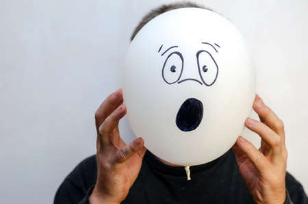 A man is holding a balloon with his face painted on it. The balloon is at head level. Unrecognizable personality. Emotion of surprise, indignation. Indoors.の写真素材