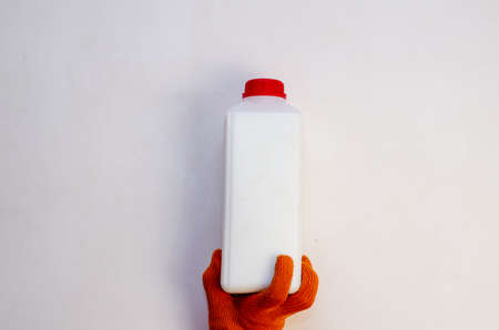 A hand in a red protective glove holds a plastic container against a light background. A mans hand with a white rectangular bottle against the wall. Paint or wall primer. Painting services. Indoors.の写真素材