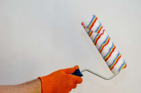 A hand in a red protective glove holds a paint roller against a light-colored wall. Mousing hand with a new hand tool. Painting services. Inside the room. Side view. Selective focus.の写真素材