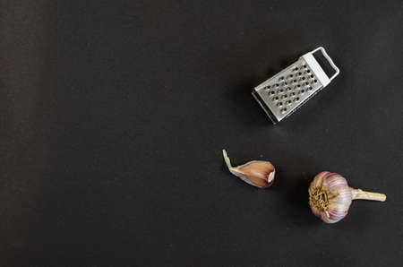 Small metal grater and a head of garlic on a dark gray background. Minimalist composition with kitchen utensils and raw garlic. Top view. Flat lay. Copy space.の写真素材