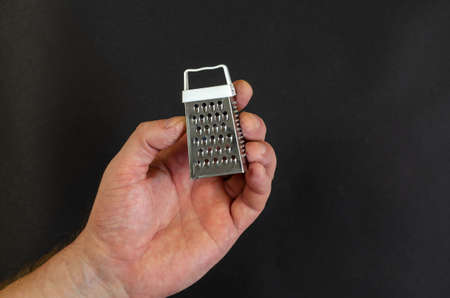 A mans hand holds a miniature metal grater against a dark gray background. Small kitchen utensil for chopping vegetables, fruit, eggs, cheese. Top view.の写真素材