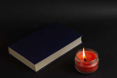 A lit candle and a closed book on a black background. Red burning candle in a clear glass jar. Bluebook in hardcover.の写真素材