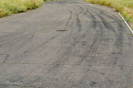 Gray asphalt with black tire tracks. An old country road with cracks. Green grass on the side of the highway. Curved wheel tracks in a bend in the driveway.の写真素材