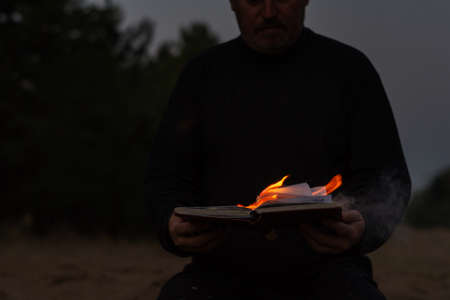 A grown man is holding a burning diary. Open notebook with tongues of bright flame. Twilight. Person sitting on the sand. Defocus, blur, noise, grain effect.の写真素材