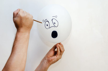 A balloon with a face painted on it and a rusty nail. Man holding white balloon and large sharp nail. Hand drawn emotion of surprise, outrage. Indoors.の写真素材