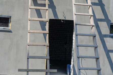 Two wooden stepladders leaned against a gray building. Plastered building with an empty doorway and window. Construction industry. Architecture.の写真素材