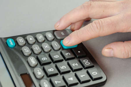 The mans index finger presses the green plus key of the calculator. The businessman performs mathematical calculations on the electronic device. Indoors. Selective focus.の写真素材