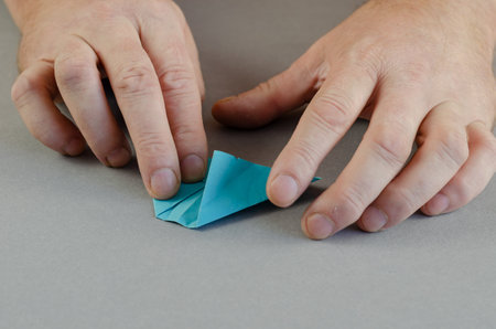 Two male hands fold a paper airplane. Blue sheet of paper. Gray background. Close-up. Selective focus.の写真素材