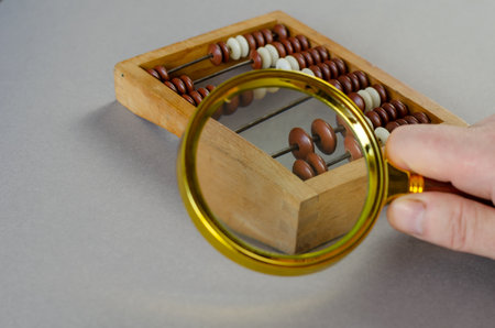 Man holds magnifying glass over small wooden abacus. Large loupe in yellow frame. Gray background. Inside the room. Close-up. Selective focus.の写真素材