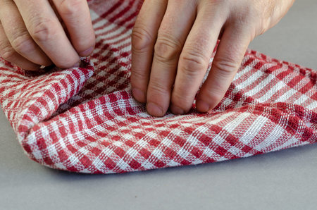A man folds a plaid napkin. Red and white cotton kitchen towel against a gray background. Close-up. Selective focus.の写真素材