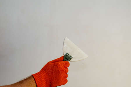 A hand in a red protective glove holds a white rubber trowel against the wall. A man with a hand-held wall putty tool. Inside the room. Side view.の写真素材