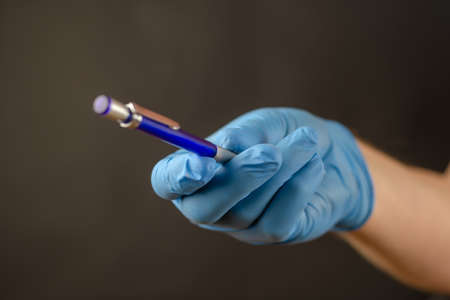 Defocused shot of doctors hand in blue medical glove with pencil. Hand is holding mechanical pencil against black background. Selective focus. Close-up. Blurred, defocus, noise, grain effectの写真素材