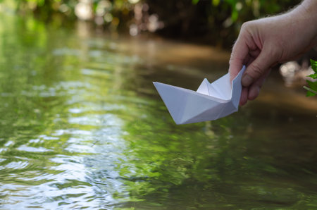 A hand lowers a white paper boat on the water. Man holds the homemade boat Shooting at water level. Selective focus. Blurred motion.の写真素材