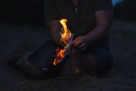 A woman holds a burning piece of paper in her hand. A grown woman sits alone on the beach at night. A torn sheet of paper from a diary with flames. Defocus, blur, noise, grain effect.の写真素材