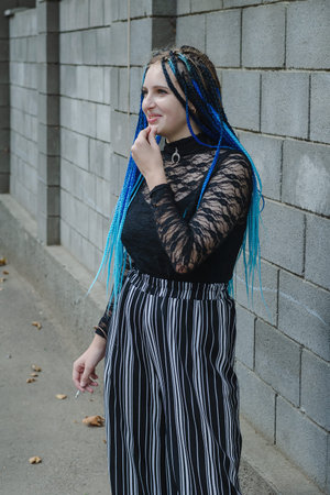 Length portrait young woman against gray stone wall. A lovely woman with long blue pigtails smokes a...