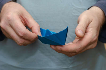 Two Hands holding a paper boat. Adult male with a blue children's boat. Close-up. Inside the room. Selective focus.の写真素材